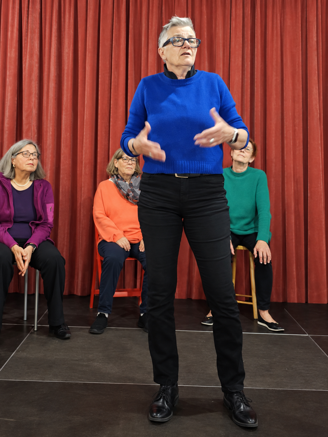 Theaterszene aus «Brücken»: Eine Darstellerin im blauen Pullover steht vor einem roten Vorhang, dahinter sitzen drei weitere Darstellerinnen.